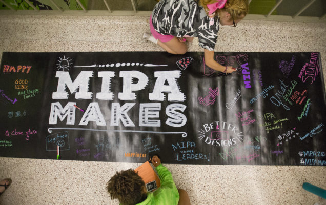 Two students sit on the floor adding text to a banner that says "MIPA Makes"
