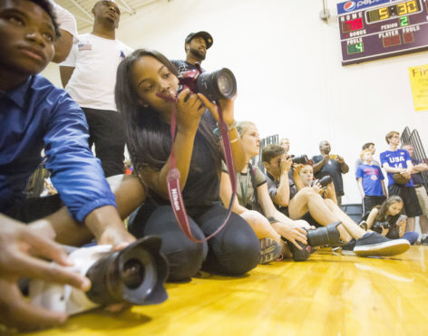 Student photographers sit and kneel on the edge of a basketball court as they watch and take photos.