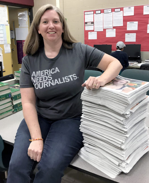 Cammie Hall sits next to a stack of newspapers wearing an America Needs Journalists shirt
